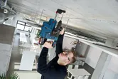En person iført sikkerhedsudstyr borer ind i et betonloft i et moderne arbejdsområde.