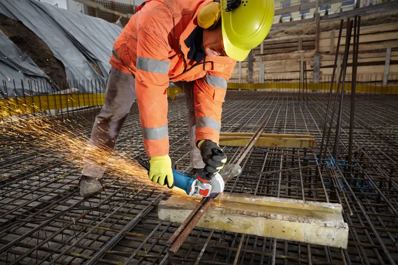 En arbejder iført sikkerhedsudstyr skærer armeringsjern på en byggeplads med en vinkelsliber.
