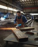En person iført sikkerhedsudstyr sliber en stor metalbjælke i et industriværksted.