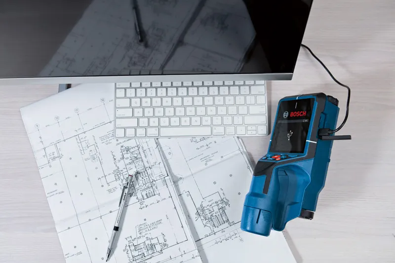 Byggedetektor forbundet til en computer ved siden af arkitekttegninger og et tastatur.