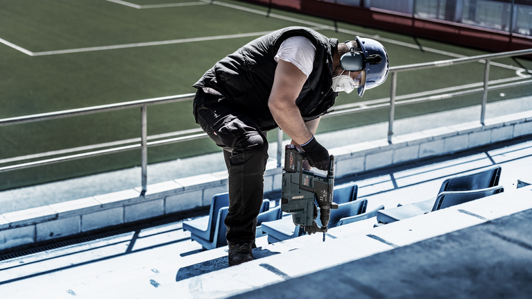 En person iført sikkerhedsudstyr borer sig ind i et betontrin på et stadion.