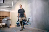En person bærer en batteridrevet geringssav og står på en moderne byggeplads.