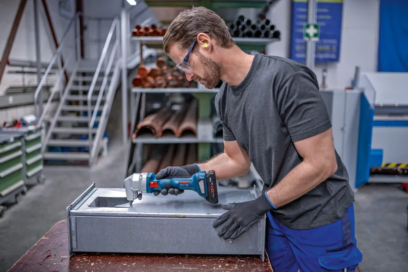 En person iført sikkerhedsudstyr trimmer metalplader med en batteridrevet nibbler.