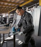 En person iført sikkerhedsudstyr saver metalplader med en batteridrevet stiksav i et værksted.