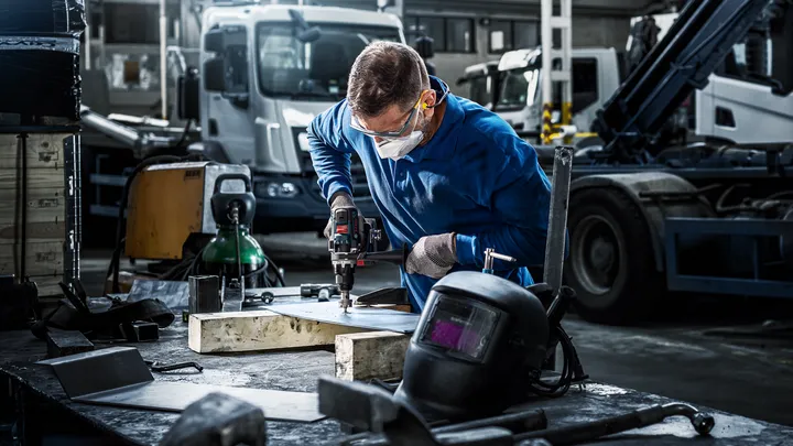Arbejder iført sikkerhedsudstyr borer metal på en arbejdsbænk i en industrigarage.