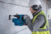 En person iført sikkerhedsudstyr borer ind i en betonvæg med en borehammer.