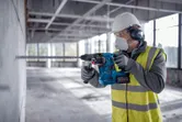 Person iført sikkerhedsudstyr borer ind i en betonvæg med en borehammer.