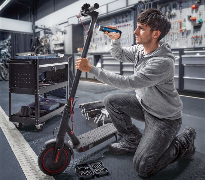 En person reparerer en elektrisk scooter med en batteridrevet skruetrækker i et værksted.