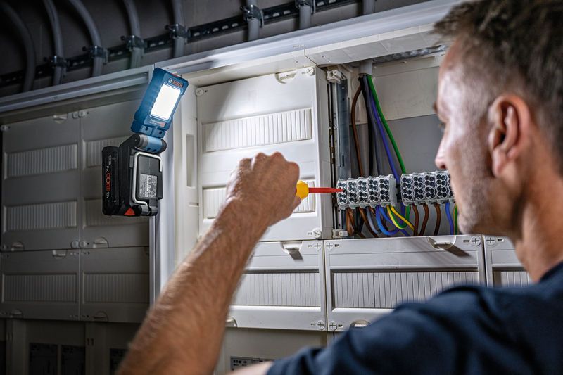 En person bruger en skruetrækker på et elektrisk panel, og en ledningsfri lampe er fastgjort til belysning.