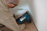 En person iført sikkerhedsudstyr bruger en batteridrevet træsømpistol til at fastgøre gulvlister.