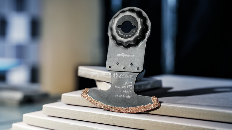 Oscillerende værktøjsklinge med hårdmetalkant hviler på stablede keramiske fliser.