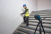 En person iført sikkerhedsudstyr markerer en væg på en trappe med et lasernivelleringsværktøj.