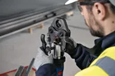 En person iført sikkerhedsudstyr betjener et batteridrevet presseværktøj på en metalfitting.