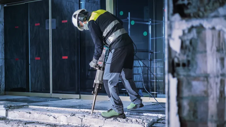 Person iført sikkerhedsudstyr bryder beton med en nedrivningshammer.