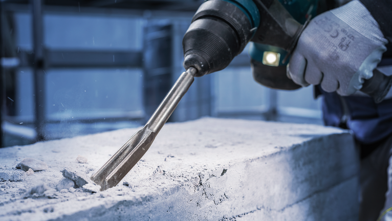 Person iført sikkerhedsudstyr mejsler beton med en borehammer.