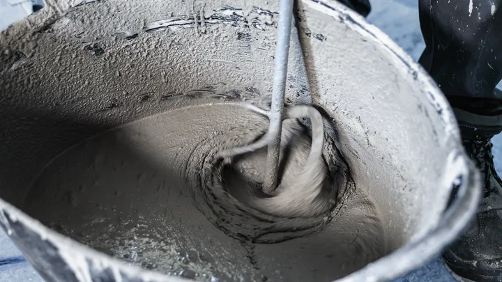 En person iført sikkerhedsudstyr blander cement med et elværktøj i en spand.