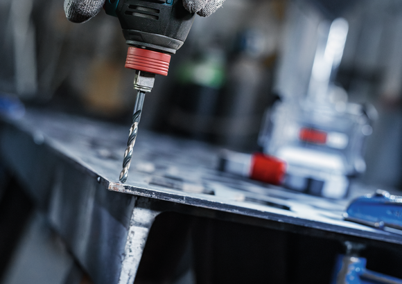 En person iført sikkerhedsudstyr borer et hul i en metalplade.