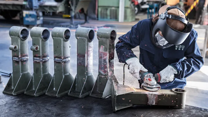 En person iført sikkerhedsudstyr sliber metaldele og producerer gnister i et værksted.