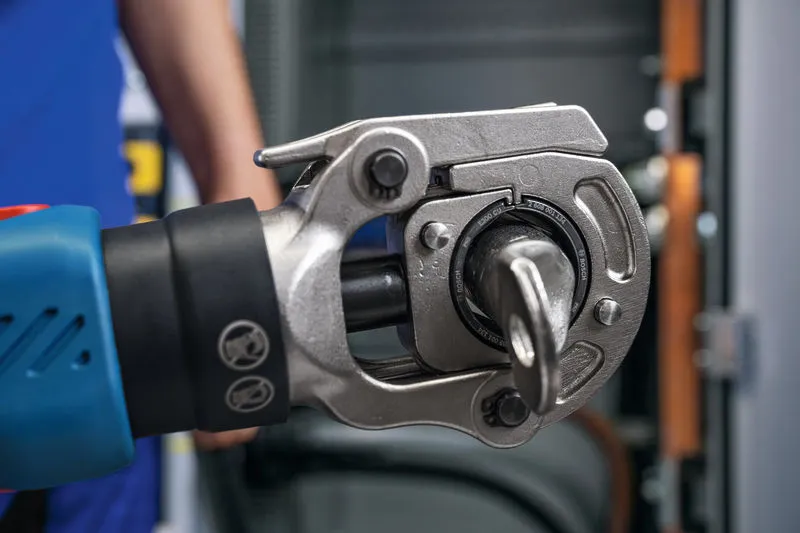 En person iført sikkerhedsudstyr betjener en batteridrevet hydraulisk krympeværktøj foran et panel.