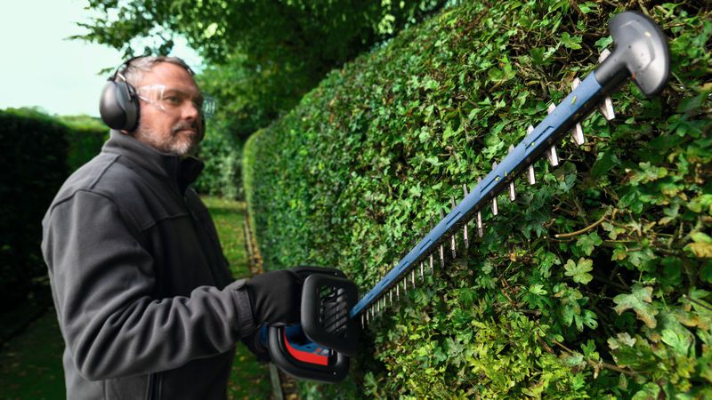 En person iført sikkerhedsudstyr trimmer en hæk med en batteridrevet hækkeklipper.