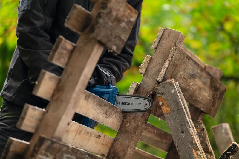 En person iført sikkerhedsudstyr saver træplanker med en batteridrevet beskæresav.