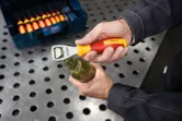 En person åbner en glasflaske med en gul og rød flaskeåbner ved en metalarbejdsbænk.