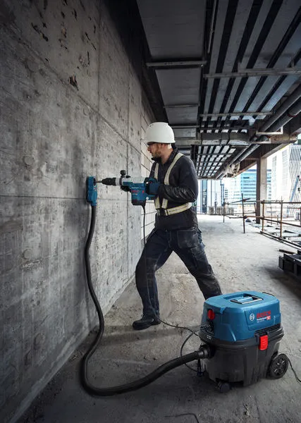 En person iført sikkerhedsudstyr borer i en betonvæg med en våd- og tørstøvsuger.
