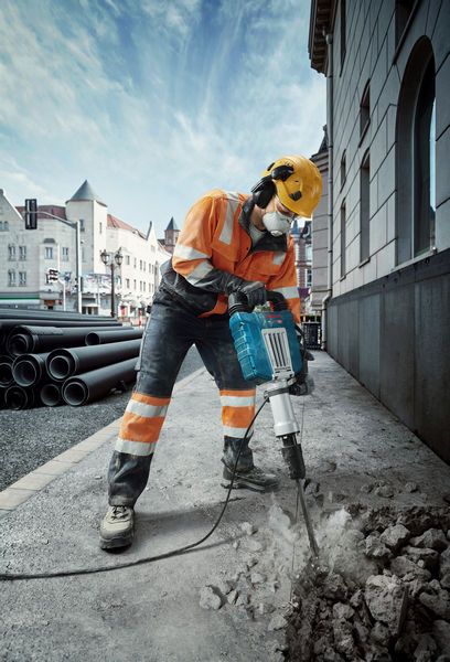 En person iført sikkerhedsudstyr bryder beton ned på et fortov med en hammer.