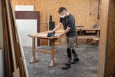 En person iført sikkerhedsudstyr sliber en trædør på en arbejdsbænk i et værksted.