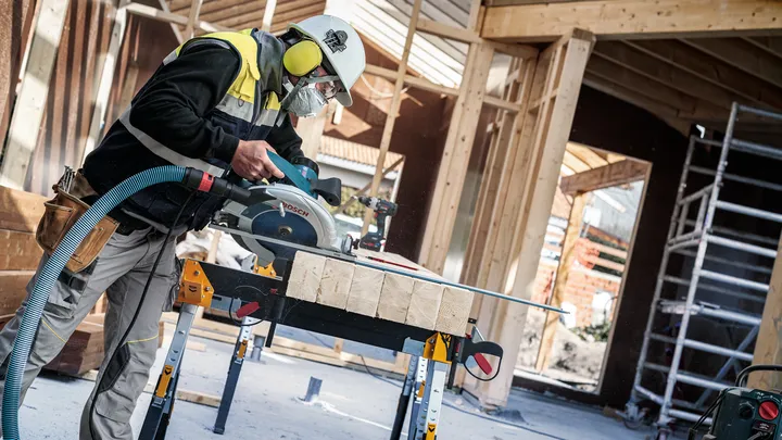 En person iført sikkerhedsudstyr saver træplanker med en rundsav på en byggeplads.