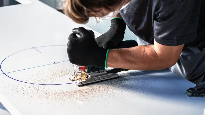 En person med sikkerhedsudstyr bruger en stiksav til at skære en kurve i en markeret bræt.