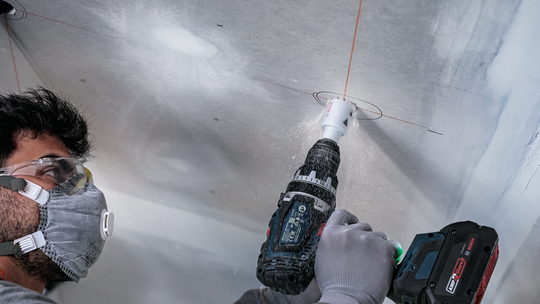 En person iført sikkerhedsudstyr borer et cirkulært hul i et loftspanel.