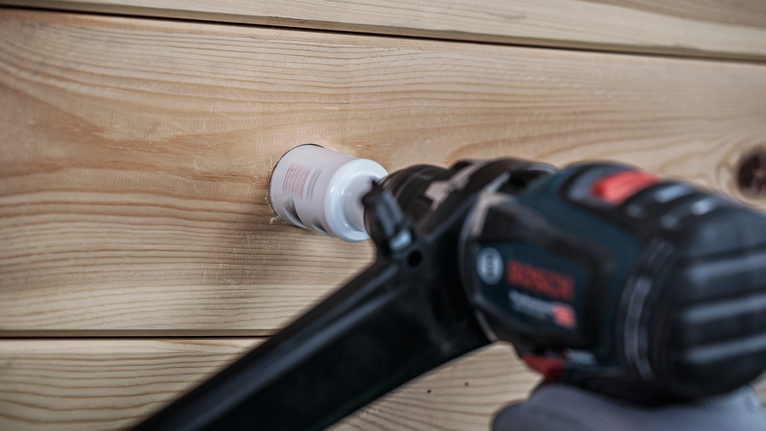 En person iført sikkerhedsudstyr borer et hul i træbrædder med en hulsav.