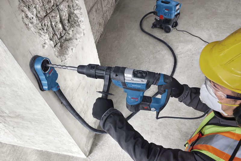 En person iført sikkerhedsudstyr borer i beton med en borehammer og støvudsuger.