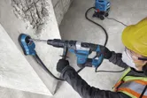 En person iført sikkerhedsudstyr borer i beton med en borehammer og støvudsuger.