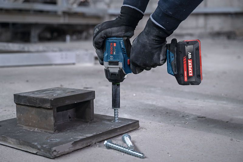 En person med sikkerhedsudstyr bruger en batteridrevet slagnøgle til at drive en bolt ind i beton.