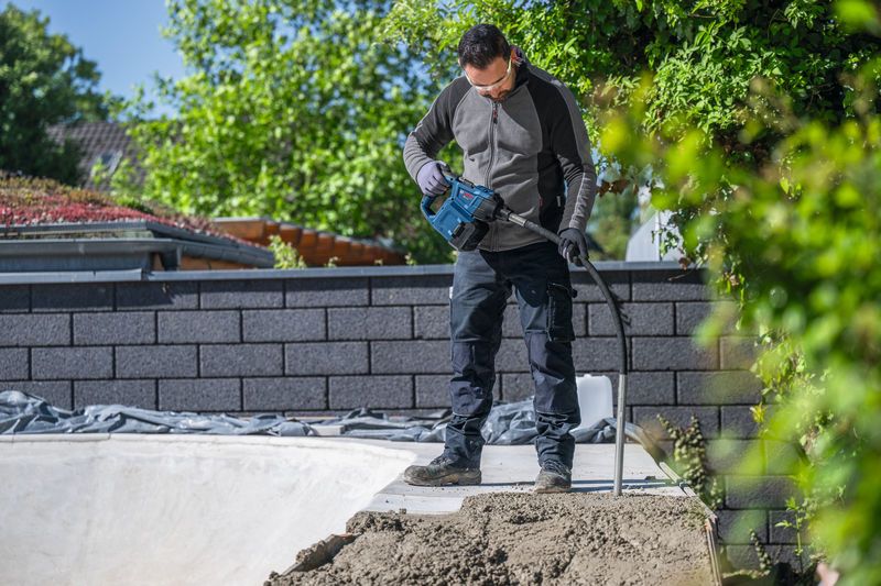 En person iført sikkerhedsudstyr bruger et elværktøj til at komprimere beton udendørs.