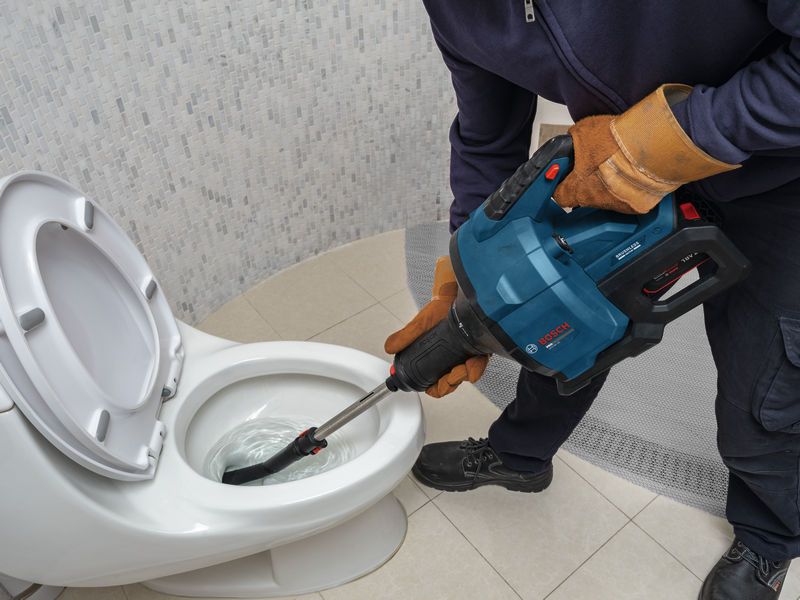 En person med sikkerhedsudstyr bruger et elværktøj til at rense et toilet.