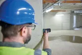 En person iført sikkerhedsudstyr bruger en lasermåler på en betonvæg.