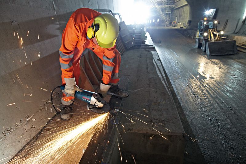 En person iført sikkerhedsudstyr sliber beton med en vinkelsliber i en tunnel.