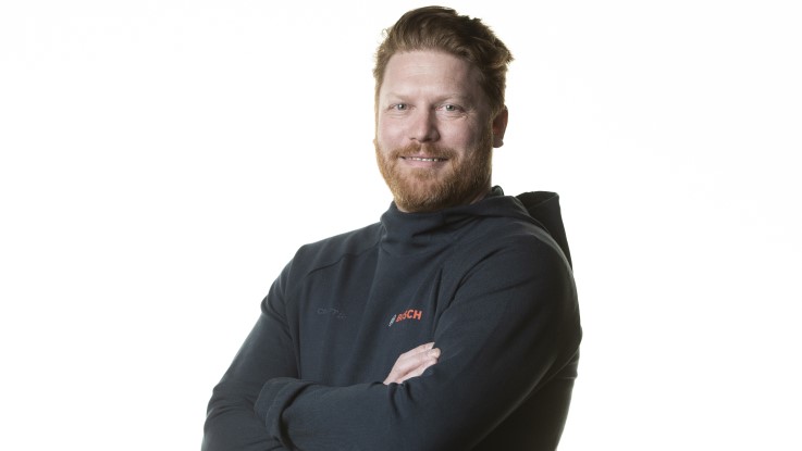 En mand med skæg iført sort trøje står med armene krydset og smiler.