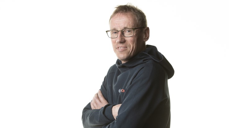 En mand i briller står med armene over kors og smiler foran en hvid baggrund.