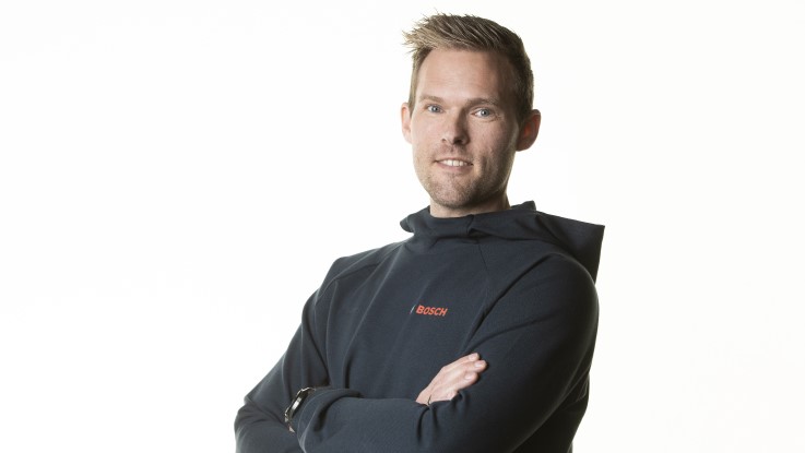 En mand med kort hår står med armene kors og smiler, iført en mørk hættetrøje.