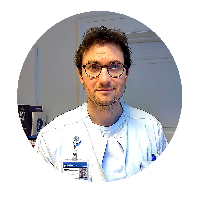 En mand i hvid lægefrakke smiler foran en hvid væg med udstyr.