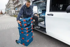 شخص يقوم بتفريغ صناديق أدوات زرقاء مكدسة من شاحنة في موقع بناء.
