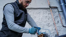 شخص يرتدي معدات السلامة يقطع أنبوبًا معدنيًا بمنشار يدوي.