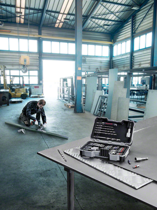 Person wearing safety equipment measures a metal sheet, with a drill bit set on a workbench.