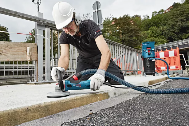 A person wearing safety equipment grinds a concrete curb with a connected dust extractor.