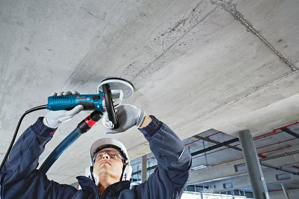 A person wearing safety equipment grinds a concrete ceiling with a power tool.