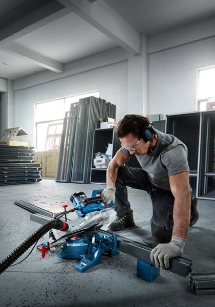 A person wearing safety equipment uses a miter saw to cut a metal beam in a workshop.